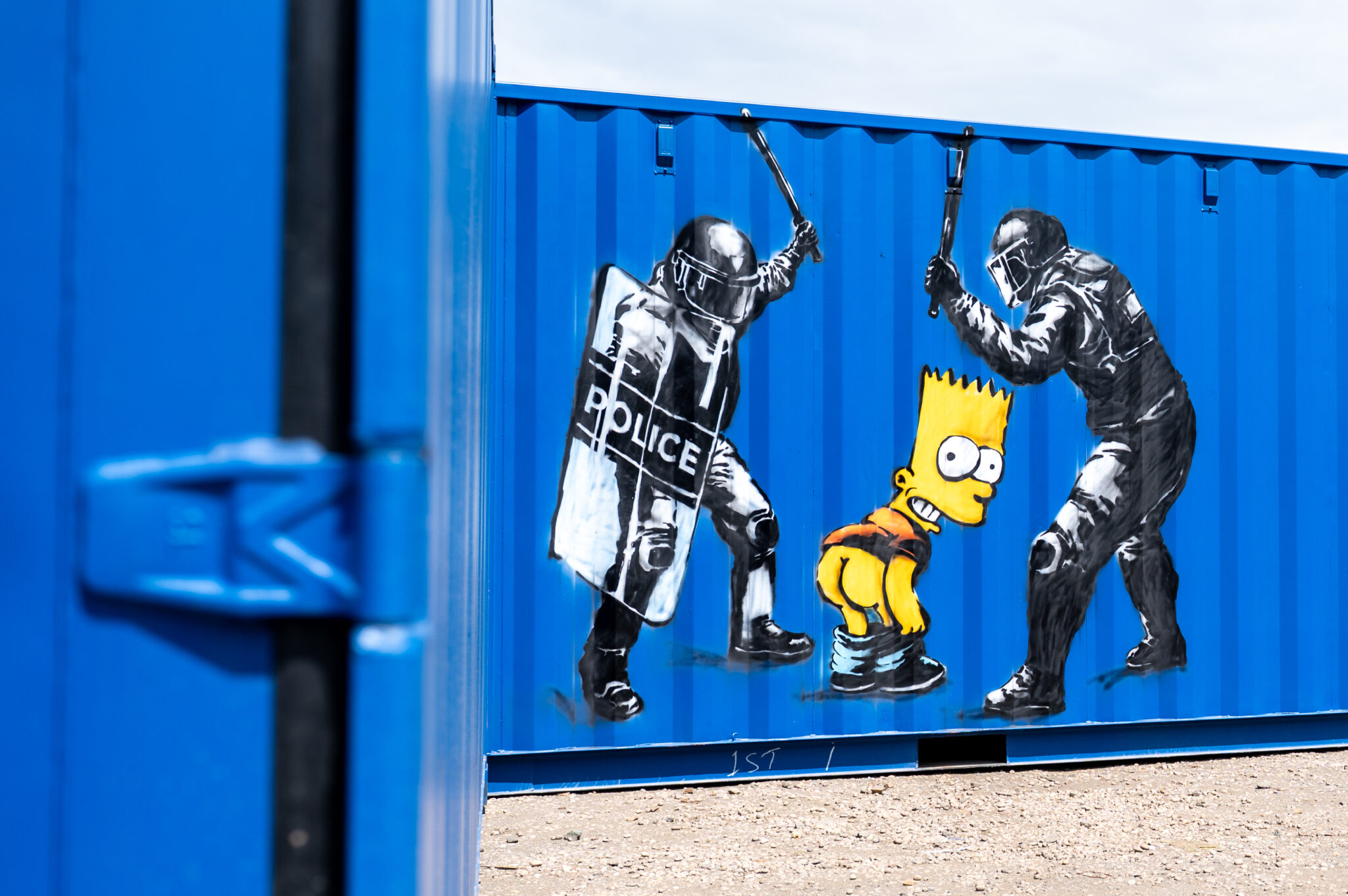 Street art mural by The Rebel Bear titled "Moony," showing Bart Simpson mooning riot police, painted on a blue BOXXS container in Linlithgow at The Rebel Bear Linlithgow Exhibition.